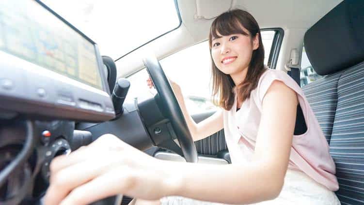 ผู้หญิงคนหนึ่งนั่งอยู่ในที่นั่งคนขับของรถยนต์ ยิ้มให้กล้องขณะหมุนกุญแจสตาร์ทและปรับการควบคุมต่างๆ.
