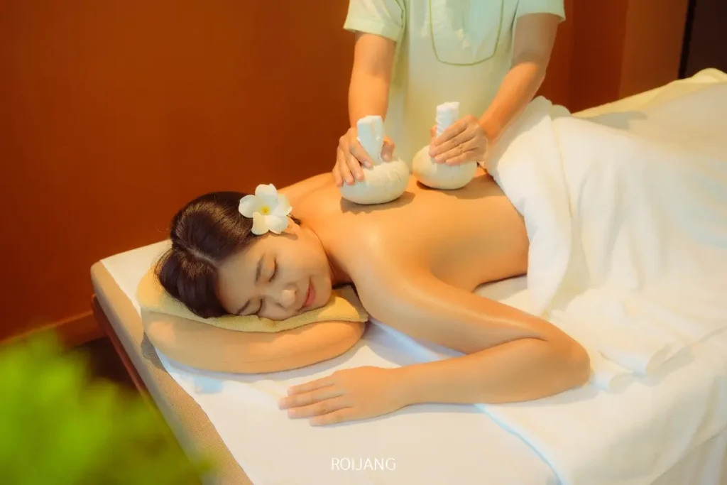 ผู้หญิงคนหนึ่งนอนคว่ำบนเตียงนวด โดยมีผ้าขนหนูคลุมส่วนล่างของร่างกายไว้ ขณะที่นักบำบัดที่ร้านสปาราตนาใช้สมุนไพรประคบอุ่นประคบลงบนหลังของเธอ.