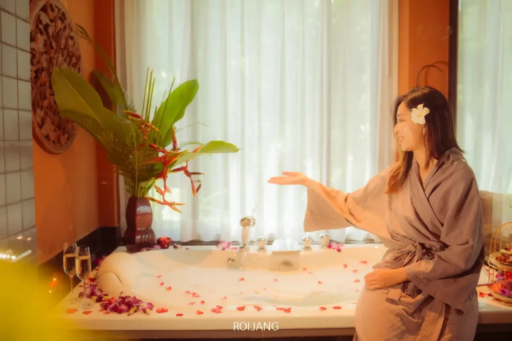 หญิงสาวในชุดคลุมอาบน้ำนั่งอยู่ข้างอ่างอาบน้ำที่เต็มไปด้วยฟองสบู่และกลีบกุหลาบที่ Ratana Spa พร้อมท่าทางเชื้อเชิญให้เข้าไปใกล้ พืชพรรณเขียวชอุ่มและไวน์สองแก้วที่อยู่ใกล้ๆ ช่วยเสริมบรรยากาศที่เงียบสงบและหรูหรา.