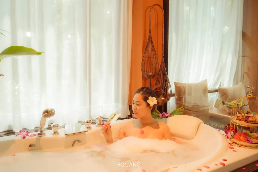 ผู้หญิงคนหนึ่งกำลังผ่อนคลายในอ่างอาบน้ำที่เต็มไปด้วยฟองสบู่พร้อมกลีบดอกไม้ที่ Ratana Spa โดยถือช้อนอยู่ และล้อมรอบไปด้วยต้นไม้เขียวชอุ่มและแสงไฟนุ่มนวลในห้องที่แสนสบาย.