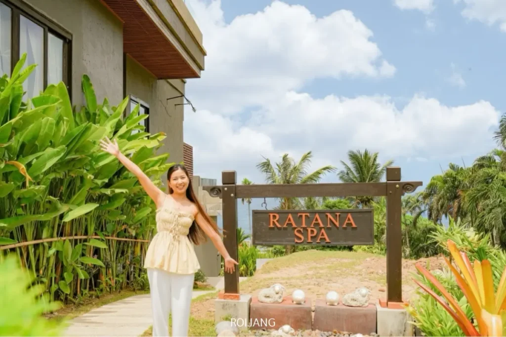 ผู้หญิงคนหนึ่งยืนยิ้มอยู่ข้างป้ายไม้ของราตนาสปา โดยมีแขนข้างหนึ่งยกขึ้น ท่ามกลางพืชเขตร้อนและท้องฟ้าแจ่มใส.