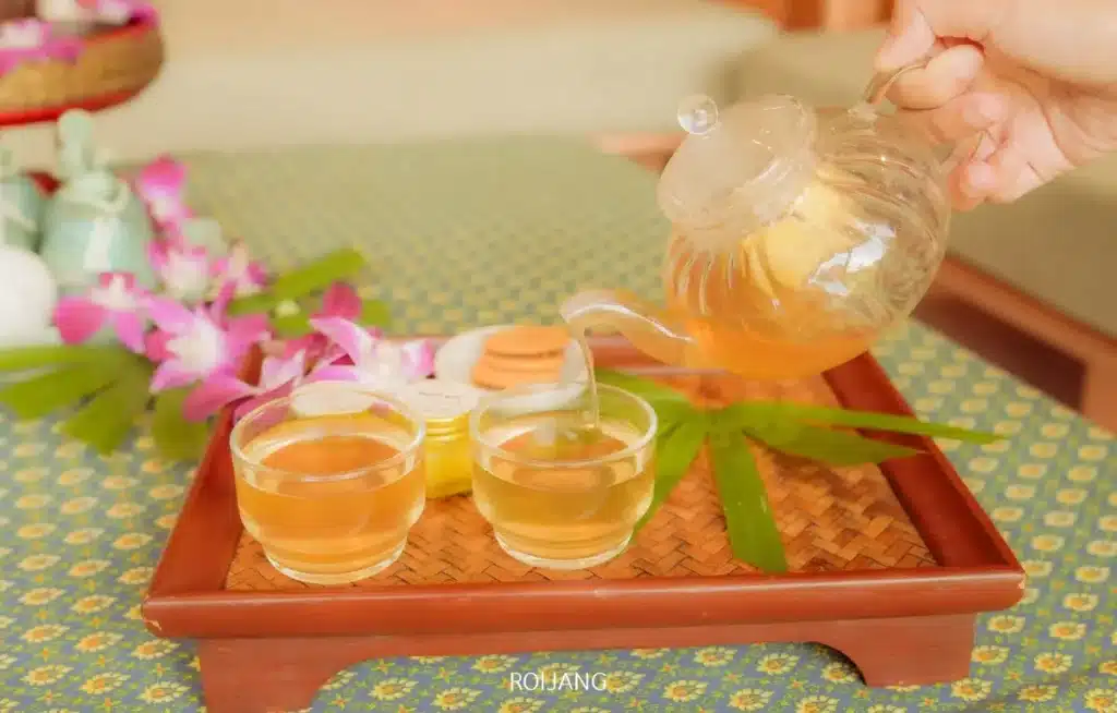 กาน้ำชาแก้วรินชาสมุนไพรลงในถ้วยแก้วบนถาดไม้ โดยมีดอกไม้และขวดโหลเล็กๆ จัดวางอยู่ข้างๆ บนผ้าปูโต๊ะลาย สร้างบรรยากาศที่สงบเงียบแบบสปารัตนะ.