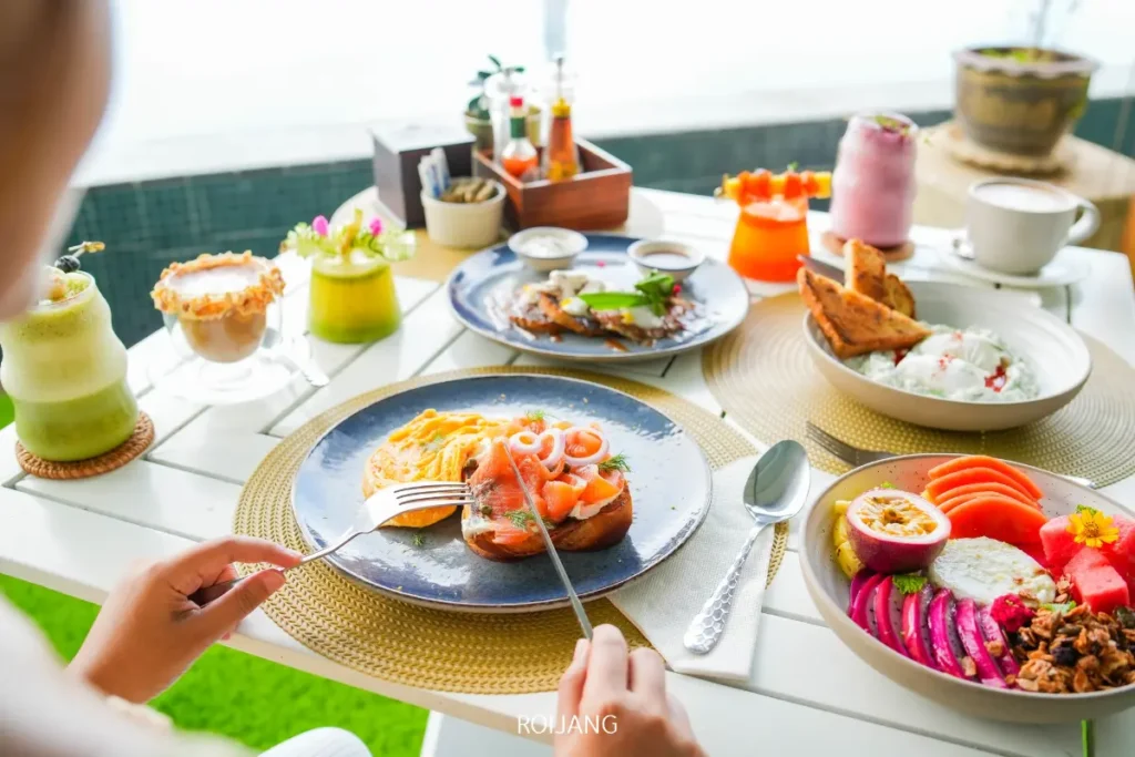 บุคคลหนึ่งนั่งอยู่ที่โต๊ะซึ่งจัดเตรียมไว้ด้วยจานอาหารเช้าสีสันสดใส เครื่องดื่ม และผลไม้ พร้อมที่จะเพลิดเพลินกับอาหารจานพิเศษที่ประกอบด้วยแซลมอนรมควันอบและไข่.