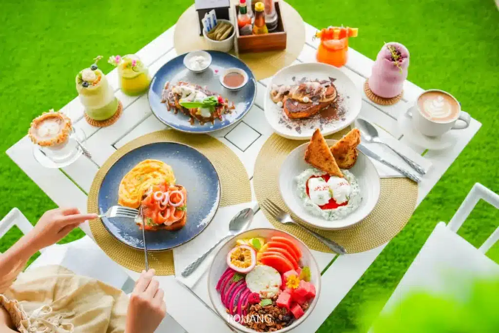 โต๊ะที่จัดเตรียมอาหารเช้าอบอย่างประณีต เครื่องดื่มหลากสีสัน และของหวานบนจานสีขาว โดยมีคนกำลังจะรับประทานอาหาร ทั้งหมดจัดวางอยู่บนพื้นผิวกลางแจ้งสีเขียวสดใส.