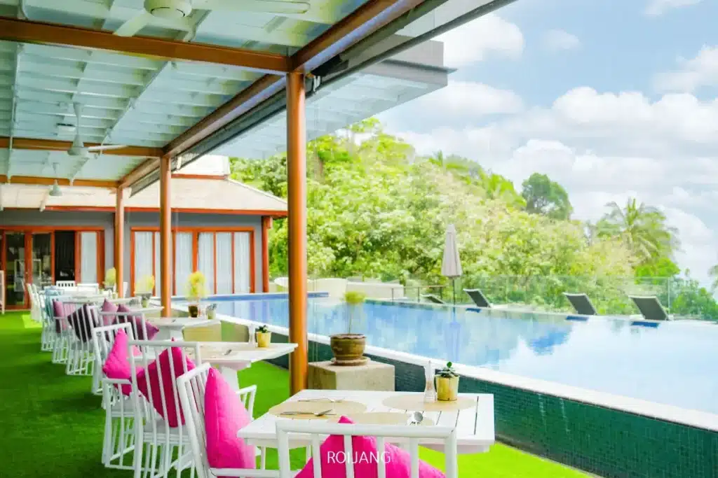 พื้นที่รับประทานอาหารกลางแจ้งพร้อมโต๊ะสีขาวและเก้าอี้เบาะสีชมพู ตั้งอยู่ข้างสระว่ายน้ำแบบอินฟินิตี้ เหมาะสำหรับการเพลิดเพลินกับขนมอบรสเลิศ ท่ามกลางธรรมชาติสีเขียวขจีและใต้เฉลียงที่มีหลังคาคลุม.