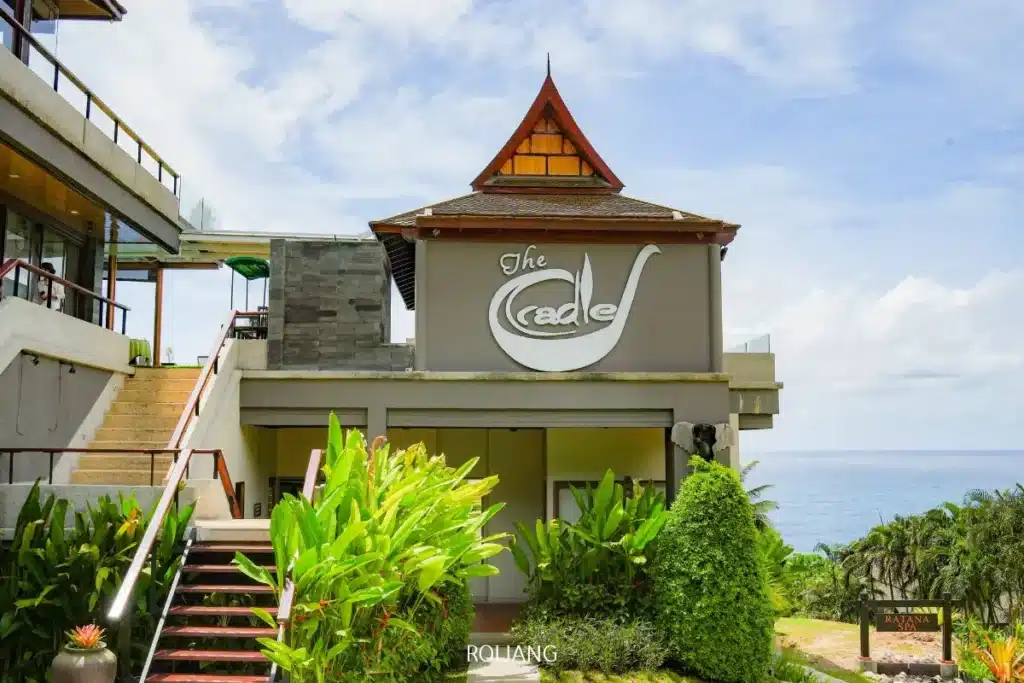 อาคารสมัยใหม่ที่มีป้ายเขียนว่า "เดอะเครเดิล" ล้อมรอบด้วยพืชเขตร้อน มองเห็นมหาสมุทรภายใต้ท้องฟ้าที่มีเมฆบางส่วน—เหมาะอย่างยิ่งสำหรับการเพลิดเพลินกับขนมอบชั้นเลิศ.