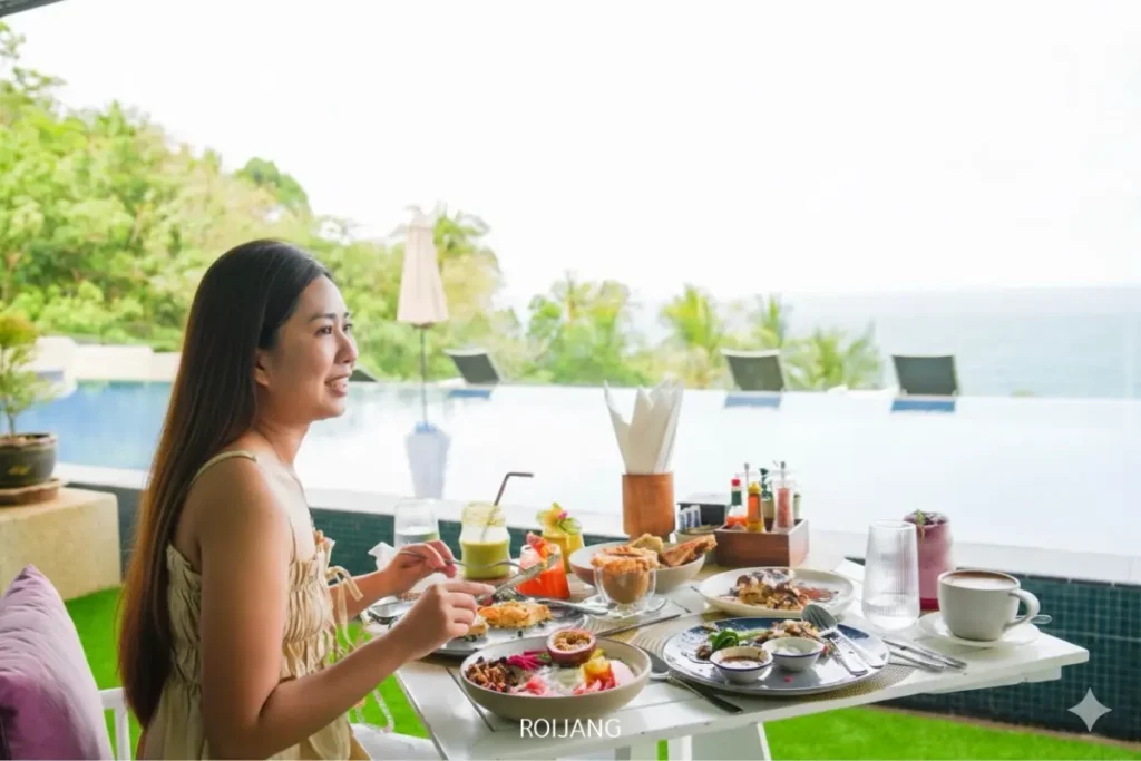 ผู้หญิงคนหนึ่งนั่งอยู่ที่โต๊ะกลางแจ้งริมสระว่ายน้ำที่ The Cradle กำลังเพลิดเพลินกับมื้ออาหารพร้อมจานหลากหลายและเครื่องดื่ม; ฉากหลังเป็นต้นไม้เขียวขจีและวิวทะเล.