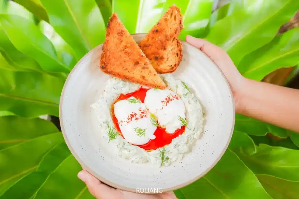 บุคคลหนึ่งถือจานที่มีขนมปังอบกรอบอย่างดี ไข่ลวกบนโยเกิร์ต ซอสสีแดง และสมุนไพร วางอยู่บนพื้นหลังของใบไม้สีเขียวขนาดใหญ่.