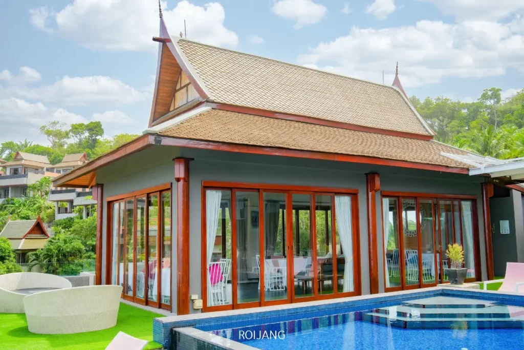 วิลล่าสมัยใหม่พร้อมหลังคาสไตล์ไทยแบบดั้งเดิม ประตูบานกระจกขนาดใหญ่ และสระว่ายน้ำด้านหน้า—ล้อมรอบด้วยที่นั่งกลางแจ้ง Fine Baked และพืชพรรณเขียวชอุ่มภายใต้ท้องฟ้าที่มีเมฆบางส่วน.