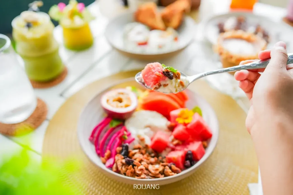 มือหนึ่งถือช้อนที่ตักโยเกิร์ตและผลไม้ไว้เหนือชามที่เต็มไปด้วยโยเกิร์ต, กรานูล่าอบกรอบ Fine Baked, แตงโม, ฝรั่ง, มะม่วงหิมพานต์, และท็อปปิ้งอื่น ๆ.