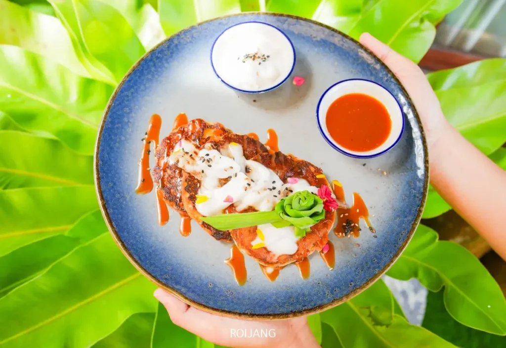 จานสีน้ำเงินที่มีเนื้อสัตว์ย่างอบละเอียดราดด้วยซอสขาว ตกแต่งด้วยผักสีเขียว และถ้วยซอสจิ้มขนาดเล็กสองใบวางอยู่บนใบเขียวขนาดใหญ่.