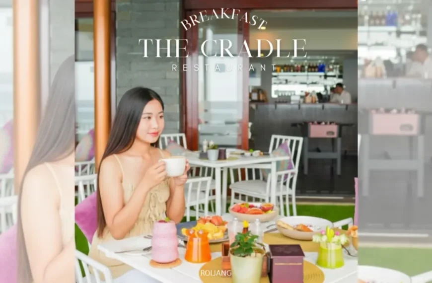ผู้หญิงคนหนึ่งนั่งอยู่ที่โต๊ะในร้านอาหารชื่อ The Cradle ล้อมรอบด้วยอาหารเช้าหลากหลายชนิดและเครื่องดื่ม เธอกำลังถือถ้วยในมือและมองไปทางข้างๆ บรรยากาศสว่างไสวและทันสมัย.
