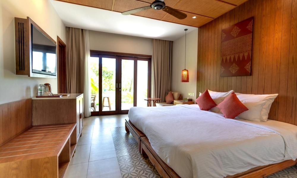 ห้องพักโรงแรมทันสมัยพร้อมเตียงขนาดใหญ่ เฟอร์นิเจอร์ไม้ ทีวีติดผนัง และวิวของลานระเบียงผ่านประตูกระจก ที่พักเชียงใหม่สไตล์ทันสมัยแห่งนี้.