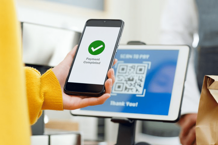 บุคคลหนึ่งถือโทรศัพท์ที่แสดงข้อความ "ชำระเงินเสร็จสมบูรณ์" อยู่ด้านหน้าหน้าจอสแกนคิวอาร์โค้ดสำหรับการชำระเงินที่เคาน์เตอร์ชำระเงิน.