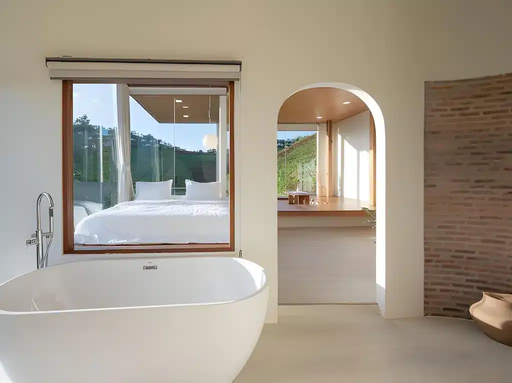 ห้องน้ำสไตล์โมเดิร์นพร้อมอ่างอาบน้ำลอยตัวสีขาว หน้าต่างบานใหญ่ที่มองเห็นห้องนอน และประตูโค้งที่นำไปสู่ห้องที่เต็มไปด้วยแสงแดดพร้อมผนังอิฐและไม้ตกแต่งอย่างสวยงาม—เหมาะสำหรับผู้ที่ต้องการที่พักเชียงใหม่ที่มีสไตล์.