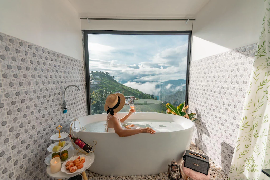 บุคคลสวมหมวกกันแดดกำลังผ่อนคลายในอ่างอาบน้ำพร้อมเครื่องดื่ม หันหน้าไปทางหน้าต่างบานใหญ่ที่มองเห็นภูเขาและเมฆ ณ โรงแรมเชียงใหม่ พร้อมของว่างและลำโพงอยู่ใกล้ๆ.