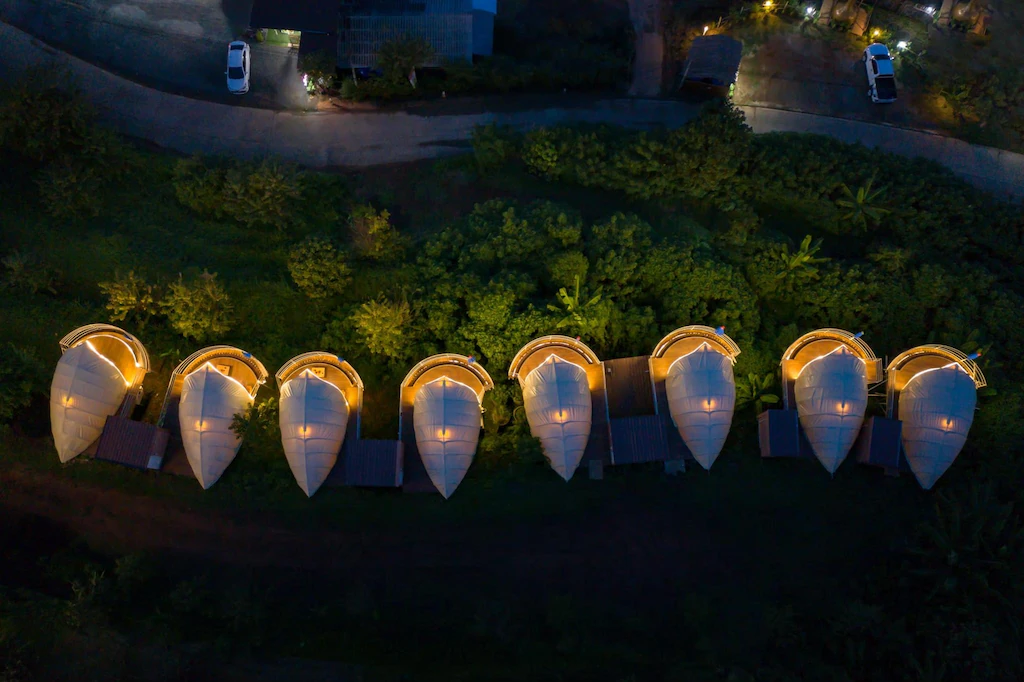 ภาพมุมสูงของกระท่อมรูปทรงเต็นท์แปดหลังที่ส่องแสงสว่างเรียงกันท่ามกลางธรรมชาติสีเขียวในยามค่ำคืน ณ รีสอร์ทเชียงใหม่สุดพิเศษ.