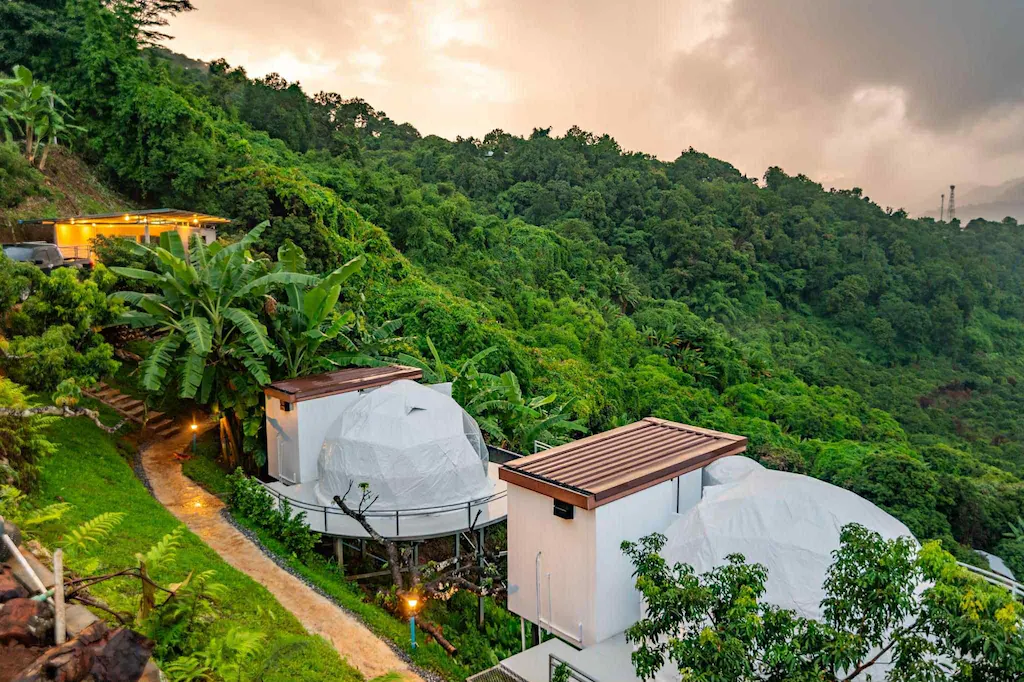 เต็นท์ทรงโดมสมัยใหม่และโครงสร้างรูปสี่เหลี่ยมผืนผ้าตั้งอยู่ท่ามกลางต้นไม้เขียวชอุ่มบนเนินเขาที่รีสอร์ทเชียงใหม่ โดยมีเส้นทางคดเคี้ยวผ่านทิวทัศน์ใต้ท้องฟ้าที่มีเมฆครึ้ม.