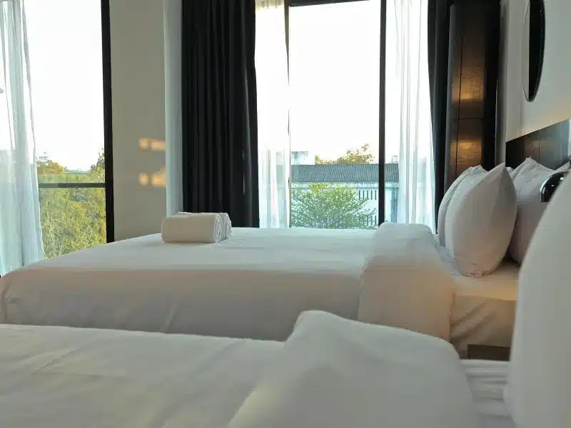 ห้องพักในโรงแรมที่เชียงใหม่มีเตียงสองเตียงที่จัดไว้อย่างเรียบร้อยพร้อมผ้าปูที่นอนและหมอนสีขาว ผ้าเช็ดตัวม้วนอยู่บนเตียงหนึ่ง ผ้าม่านขนาดใหญ่และผ้าม่านบางที่ปล่อยให้แสงแดดส่องเข้ามาอย่างนุ่มนวล.