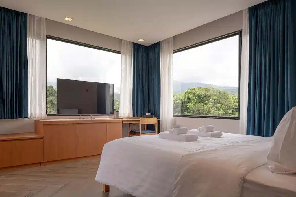 ห้องพักโรงแรมสมัยใหม่ในโรงแรมที่เชียงใหม่ พร้อมหน้าต่างมุมขนาดใหญ่ เตียงพร้อมผ้าปูที่นอนและผ้าเช็ดตัวสีขาว โต๊ะไม้และตู้ลิ้นชัก โทรทัศน์จอแบนติดตั้งตรงข้ามเตียง สามารถชมวิวต้นไม้และภูเขาได้ภายนอก.