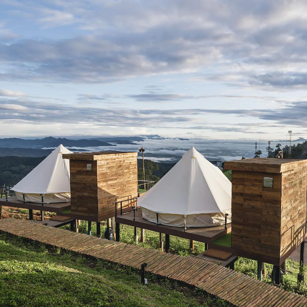 กระท่อมไม้สามหลังพร้อมเต็นท์ผ้าใบสีขาวตั้งอยู่บนเนินหญ้าที่มองเห็นภูเขาและท้องฟ้าที่มีเมฆปกคลุม—ประสบการณ์ที่พักเชียงใหม่ที่เชิญชวนสำหรับคนรักธรรมชาติ.