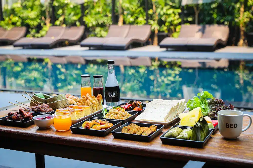 โต๊ะไม้ข้างสระว่ายน้ำที่รีสอร์ทเชียงใหม่จัดวางอาหารหลากหลายชนิด ขนม เครื่องปรุง น้ำส้ม เครื่องดื่มบรรจุขวด และแก้วกาแฟ พร้อมเก้าอี้เลานจ์และต้นไม้เขียวขจีเป็นฉากหลัง.