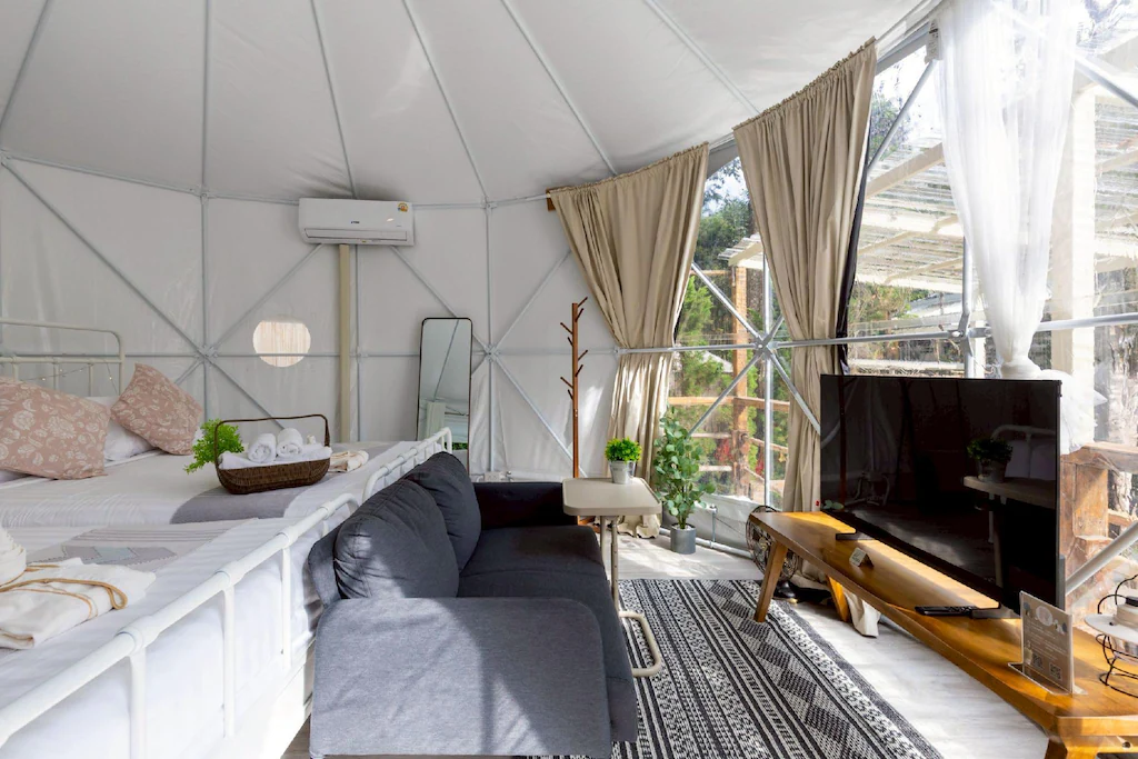 ภายในเต็นท์แกลมปิ้งทรงโดมสมัยใหม่พร้อมเตียง โซฟา โทรทัศน์ และหน้าต่างบานใหญ่พร้อมผ้าม่าน; แสงธรรมชาติส่องสว่างที่พักเชียงใหม่แห่งนี้อย่างมีเอกลักษณ์ เนื่องจากสามารถมองเห็นต้นไม้สีเขียวได้จากภายนอก.