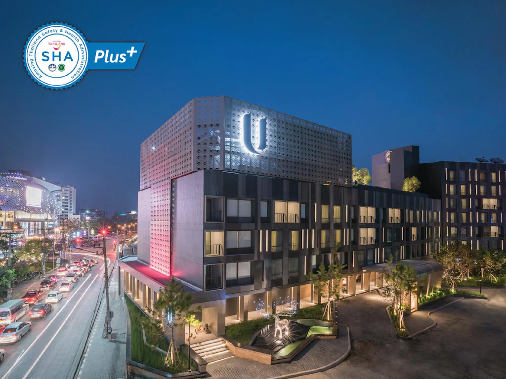 โรงแรมเชียงใหม่สมัยใหม่ในยามพลบค่ำ พร้อมโลโก้ตัว "U" ส่องแสง ถนนที่พลุกพล่านพร้อมการจราจรทางด้านซ้าย และตราสัญลักษณ์ "SHA Plus+" ที่มุมบนซ้าย.