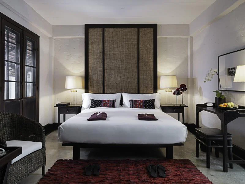 ห้องพักโรงแรมเชียงใหม่สไตล์โมเดิร์นพร้อมเตียงคู่ โต๊ะข้างเตียงและโคมไฟ เก้าอี้สาน โต๊ะทำงานพร้อมผลไม้ รองเท้าแตะบนพรมสีแดง และการตกแต่งโทนสีเรียบ—เหมาะสำหรับการเข้าพักของคุณในเชียงใหม่.