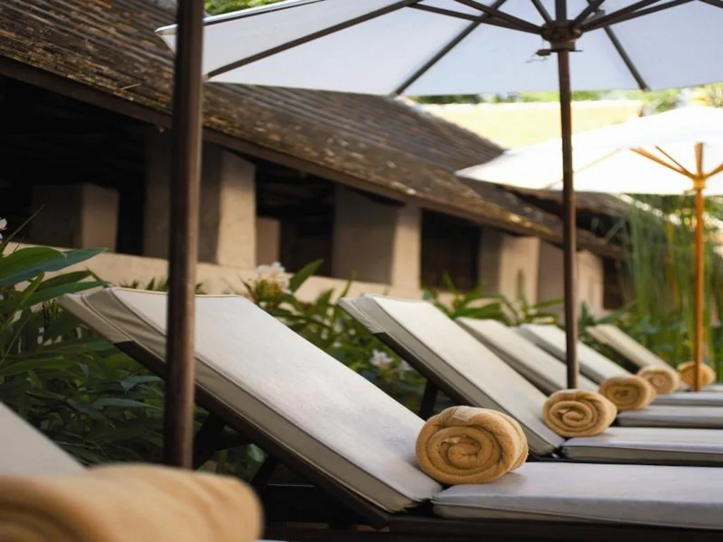 แถวของเก้าอี้เลานจ์พร้อมเบาะรองนั่งที่มีผ้าขนหนูม้วนอยู่ใต้ร่มขนาดใหญ่ข้างอาคาร ล้อมรอบด้วยพืชพรรณสีเขียวที่รีสอร์ทเชียงใหม่สุดมีสไตล์.