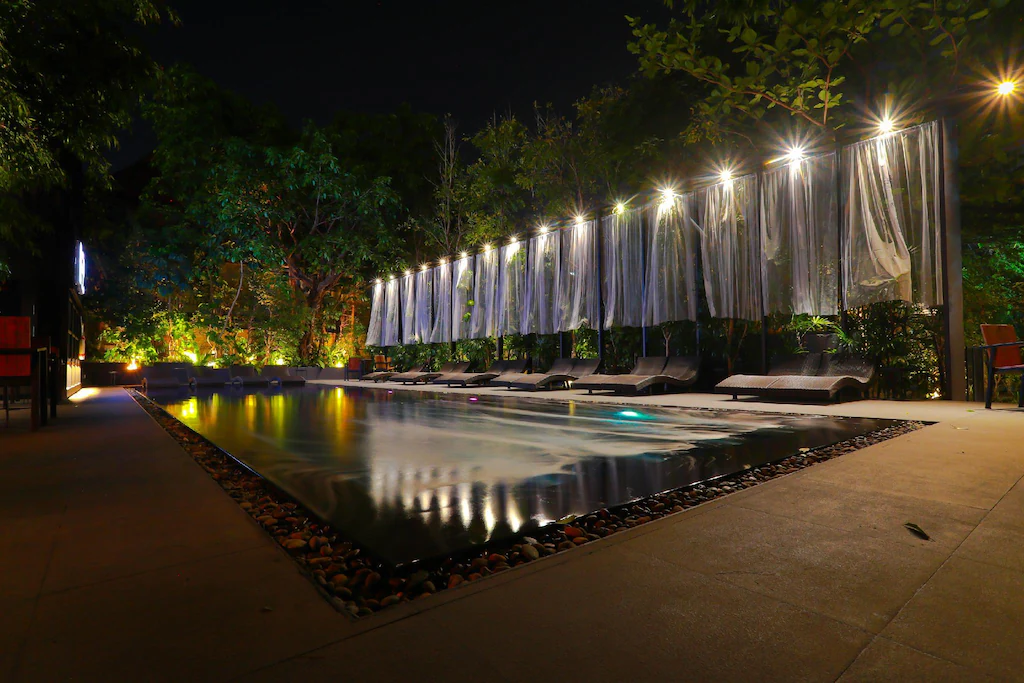 สระว่ายน้ำกลางแจ้งรูปสี่เหลี่ยมผืนผ้าในยามค่ำคืน รายล้อมไปด้วยเก้าอี้อาบแดด และประดับด้วยไฟประดับแบบสายไฟ พร้อมม่านบางเบาที่ให้ความเป็นส่วนตัวบางส่วน—เหมาะอย่างยิ่งสำหรับค่ำคืนที่ผ่อนคลายที่รีสอร์ทเชียงใหม่หรือโรงแรมในเชียงใหม่.