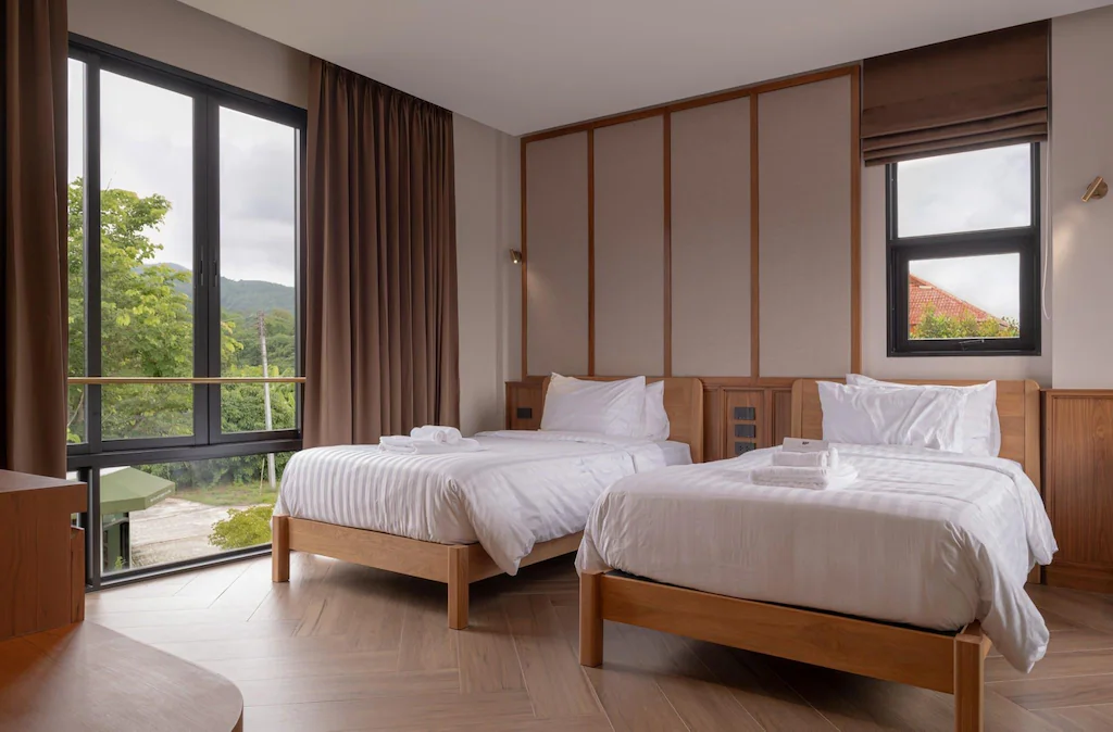 ห้องพักโรงแรมเชียงใหม่สมัยใหม่พร้อมเตียงเดี่ยวสองเตียง ตกแต่งด้วยไม้ หน้าต่างบานใหญ่พร้อมผ้าม่าน และวิวทิวทัศน์สีเขียวสดชื่นภายนอก.