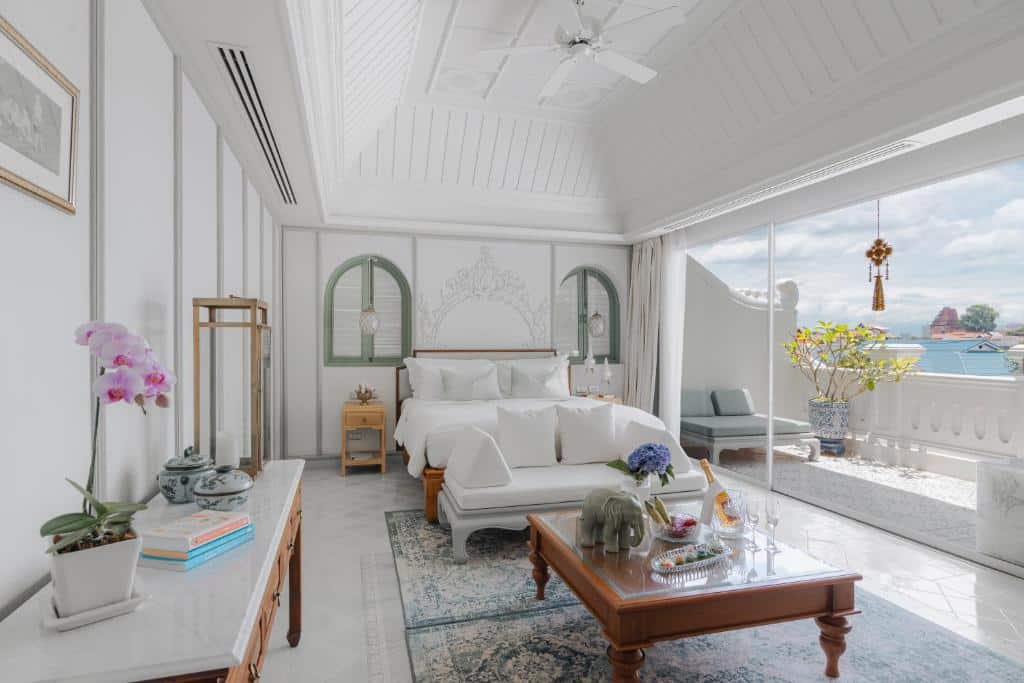 ห้องนอนสว่างไสวและหรูหราที่ โรงแรมเชียงใหม่ พร้อมผนังสีขาว เตียงใหญ่ โซฟา และโต๊ะกาแฟ; ระเบียงพร้อมต้นไม้ในกระถางและวิวทะเลสวยผ่านประตูกระจก.