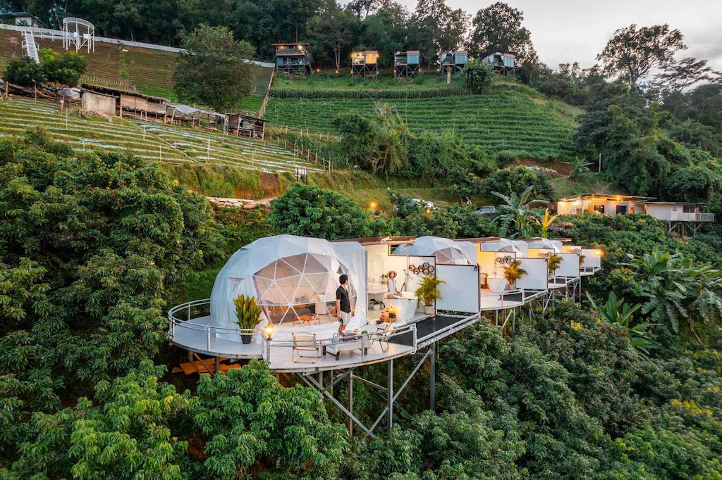 กระท่อมโดมทรงกลมบนเสาสูงสร้างบนเนินเขาที่เขียวชอุ่มล้อมรอบด้วยธรรมชาติสีเขียว โดยมีผู้คนยืนอยู่บนระเบียงหนึ่งในนั้นที่รีสอร์ทเชียงใหม่แห่งนี้.