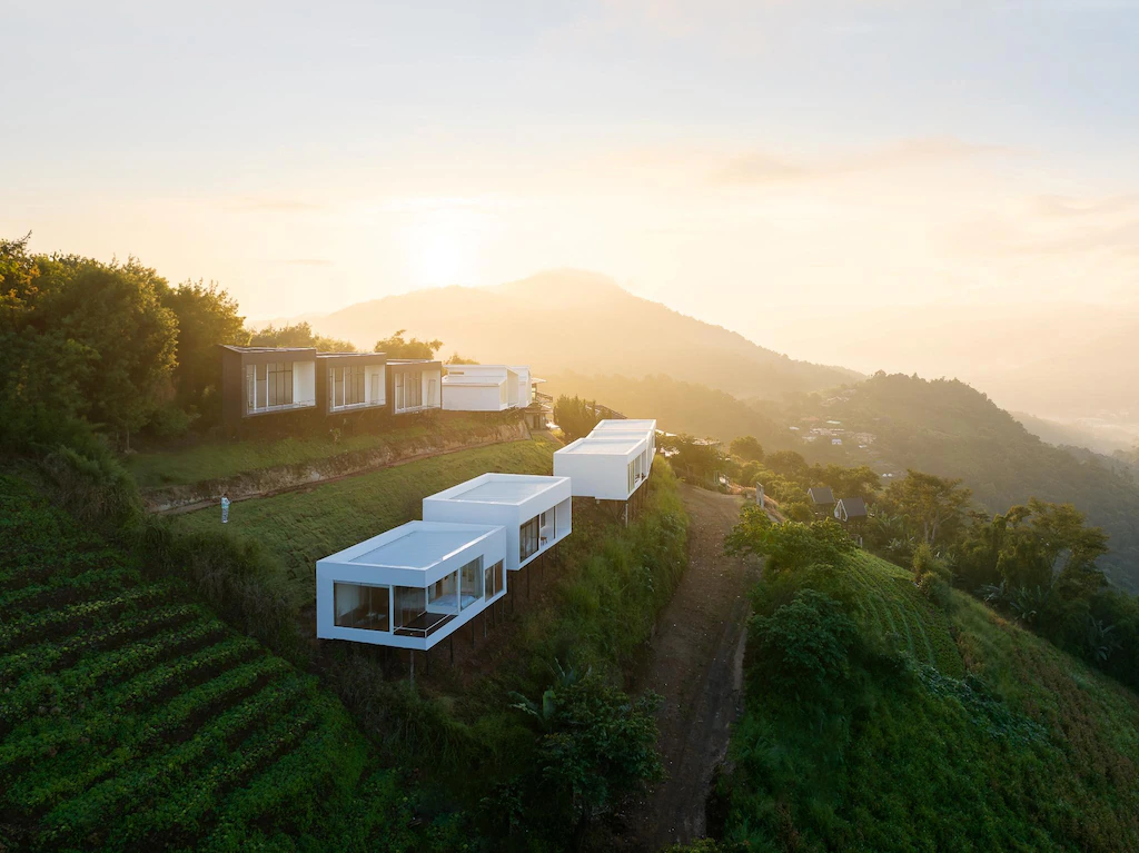 อาคารสี่เหลี่ยมผืนผ้าสีขาวสมัยใหม่ ซึ่งเป็นส่วนหนึ่งของรีสอร์ทเชียงใหม่ ถูกจัดเรียงเป็นแถวบนเนินเขาสีเขียว โดยมีพืชพันธุ์เป็นขั้นบันไดและภูเขาเป็นฉากหลังภายใต้ท้องฟ้าที่มีหมอกบางในยามพระอาทิตย์ขึ้น.