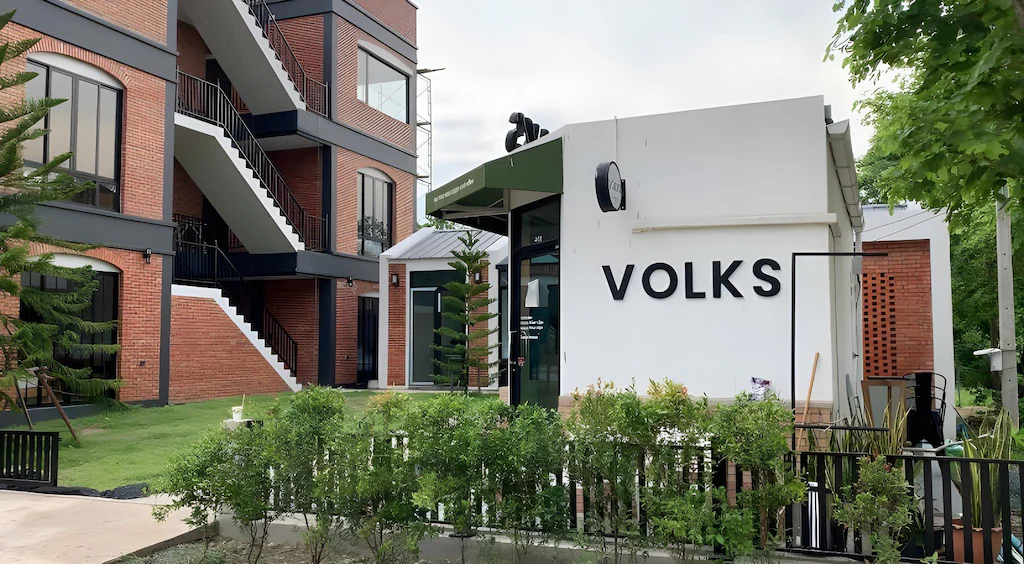 อาคารสมัยใหม่ที่มีโครงสร้างสีขาวพร้อมป้าย "VOLKS" อยู่ด้านหน้าอาคารอิฐหลายชั้น ล้อมรอบด้วยพื้นที่สีเขียว เหมาะสำหรับผู้ที่มองหาโรงแรมในเชียงใหม่หรือที่พักที่มีเอกลักษณ์ในใจกลางเมือง.