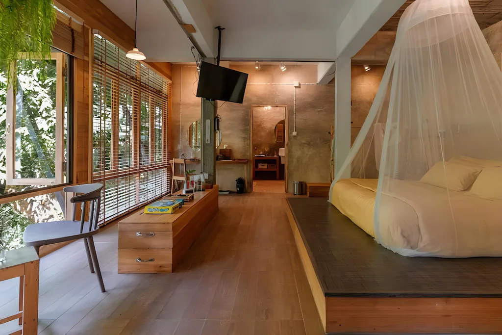 ห้องนอนกว้างขวางพร้อมเตียงหลังคา, เฟอร์นิเจอร์ไม้, หน้าต่างบานใหญ่พร้อมม่านบังตา, ทีวีติดผนัง, และวิวต้นไม้เขียวขจีภายนอก—เหมาะสำหรับการผ่อนคลายที่ โรงแรมเชียงใหม่ แห่งนี้.