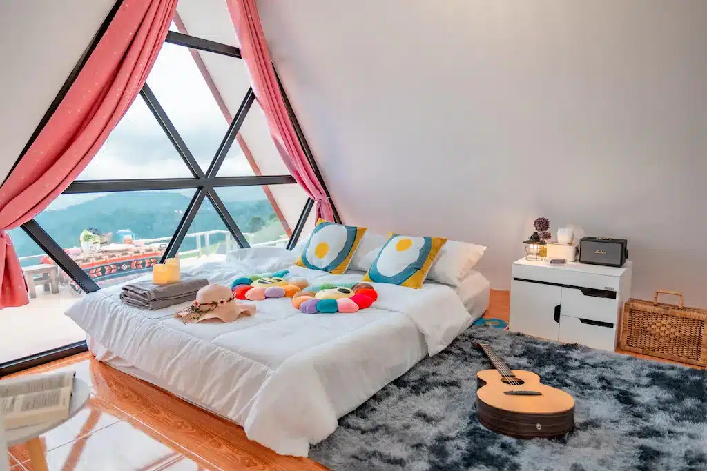 ห้องนอนที่แสนอบอุ่นในรีสอร์ทเชียงใหม่ พร้อมเตียงเตี้ย หมอนสีสันสดใส กีตาร์ และหน้าต่างสามเหลี่ยมบานใหญ่พร้อมผ้าม่านสีชมพู มองเห็นวิวทิวทัศน์ภายนอกที่สวยงาม.
