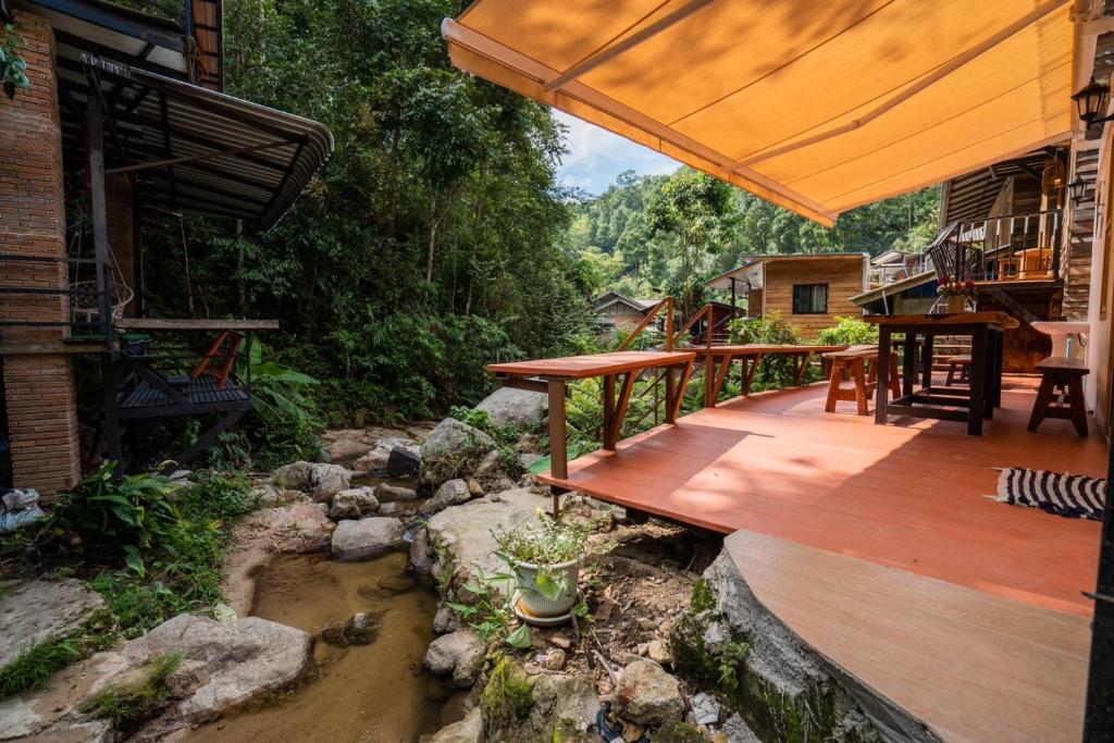 ระเบียงไม้พร้อมโต๊ะและเก้าอี้ทอดยาวออกไปเหนือลำธารหินเล็ก ๆ ล้อมรอบด้วยต้นไม้เขียวชอุ่มและบ้านไม้ใกล้เคียง—เหมาะสำหรับการพักผ่อนที่รีสอร์ทเชียงใหม่ที่มีเสน่ห์.