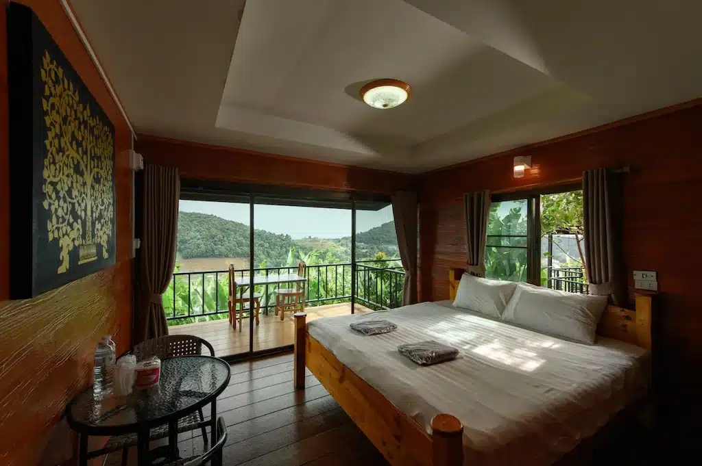 ที่พักเชียงใหม่แสนอบอุ่นพร้อมเตียงขนาดใหญ่ โต๊ะเล็กและเก้าอี้ ผ้าเช็ดตัวบนเตียง และระเบียงที่มองเห็นวิวเนินเขาสีเขียวและน้ำ.