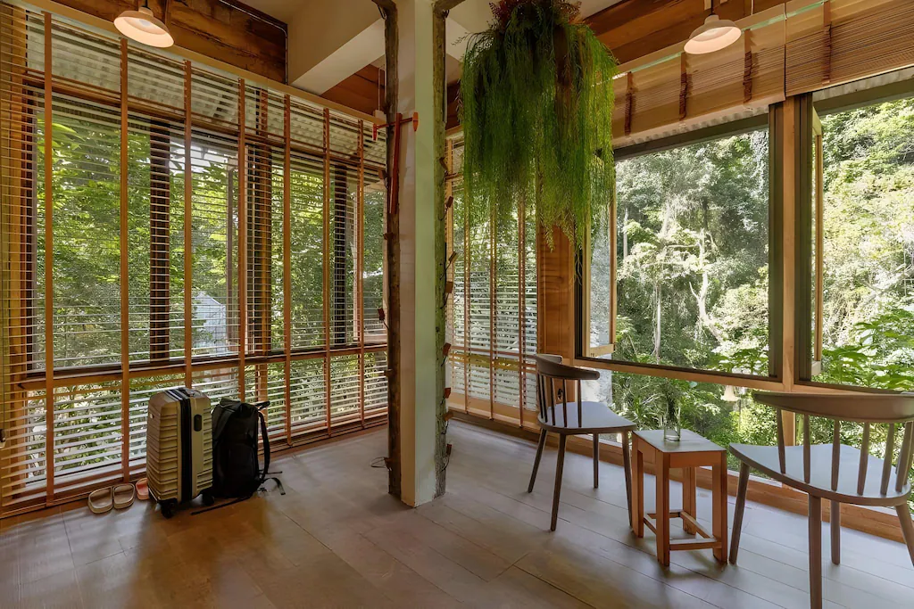 ห้องที่เต็มไปด้วยแสงแดด มีหน้าต่างบานใหญ่ เฟอร์นิเจอร์ไม้ ต้นไม้แขวน กระเป๋าเป้ และเครื่องทำความร้อนแบบพกพา มองออกไปเห็นต้นไม้เขียวชอุ่ม—เหมาะสำหรับการพักผ่อนที่ โรงแรมเชียงใหม่.