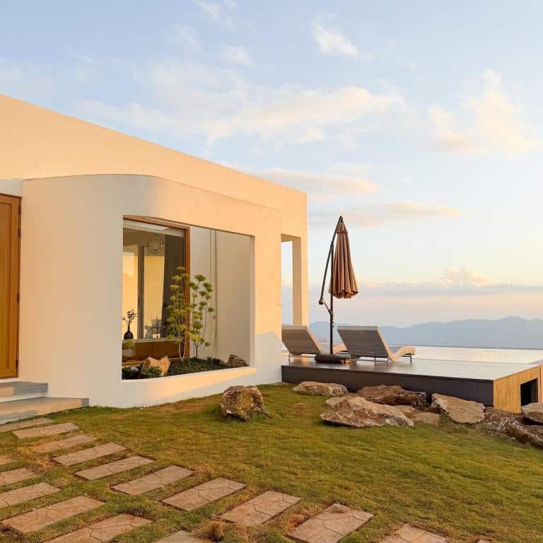 บ้านสีขาวสมัยใหม่พร้อมหน้าต่างบานใหญ่ เก้าอี้เลานจ์กลางแจ้ง ร่ม และทางเดินหินบนสนามหญ้าที่มองเห็นวิวแม่น้ำและภูเขาไกลในยามพระอาทิตย์ตก—เหมาะอย่างยิ่งสำหรับประสบการณ์การพักผ่อนที่เงียบสงบในเชียงใหม่.