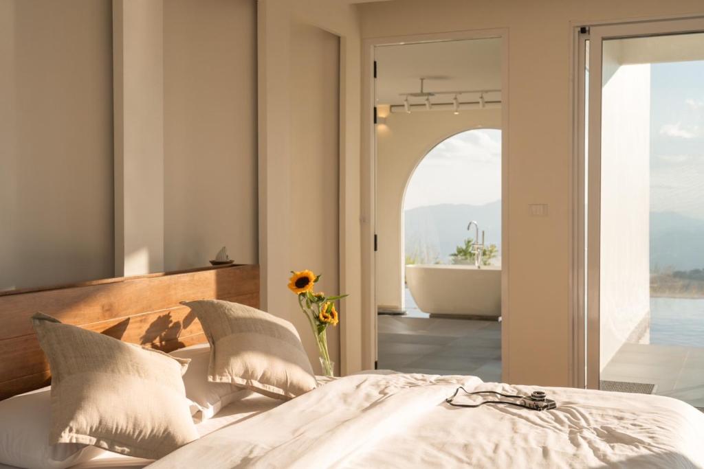 ห้องนอนที่อาบแสงแดดในรีสอร์ทเชียงใหม่ มีหัวเตียงไม้ หมอนสีเบจ ดอกทานตะวันในแจกัน และประตูเปิดไปยังห้องน้ำที่มีอ่างอาบน้ำและวิวภูเขาที่สวยงามตระการตา.