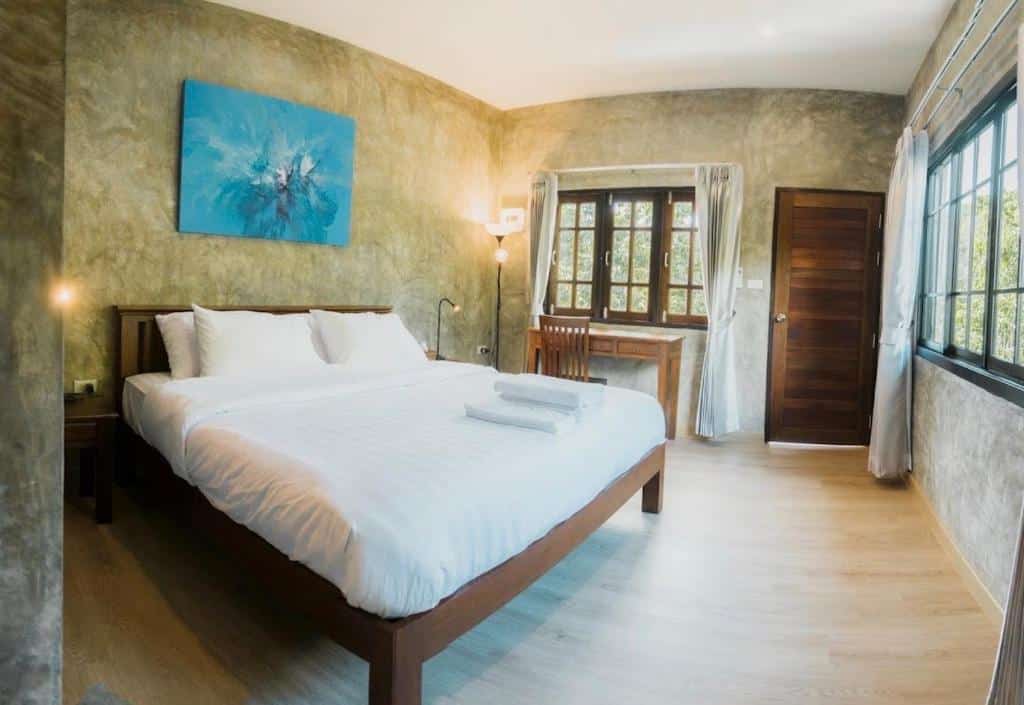 เตียงที่จัดไว้อย่างเรียบร้อยพร้อมผ้าปูที่นอนสีขาวในห้องนอนโรงแรมเชียงใหม่สไตล์โมเดิร์นที่มีพื้นไม้ โต๊ะทำงาน หน้าต่างบานใหญ่ และศิลปะผนังแบบนามธรรม.