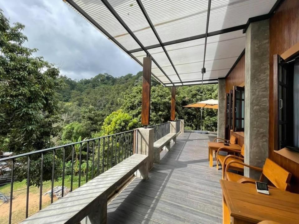 ระเบียงที่มีหลังคาคลุมที่โรงแรมเชียงใหม่แห่งหนึ่งมีเก้าอี้ไม้และโต๊ะตั้งอยู่ มองเห็นป่าเขียวชอุ่มเบื้องล่างภายใต้ท้องฟ้าที่มีเมฆบางส่วน โดยมีร่มปรากฏให้เห็นอยู่ไกลออกไป.