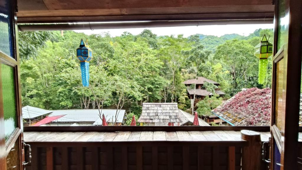 วิวจากระเบียงไม้ที่รีสอร์ทเชียงใหม่ มองเห็นหลังคาบ้าน ต้นไม้ และภูเขา พร้อมโคมไฟสีสันสดใสสองดวงแขวนอยู่ที่ชายคา.