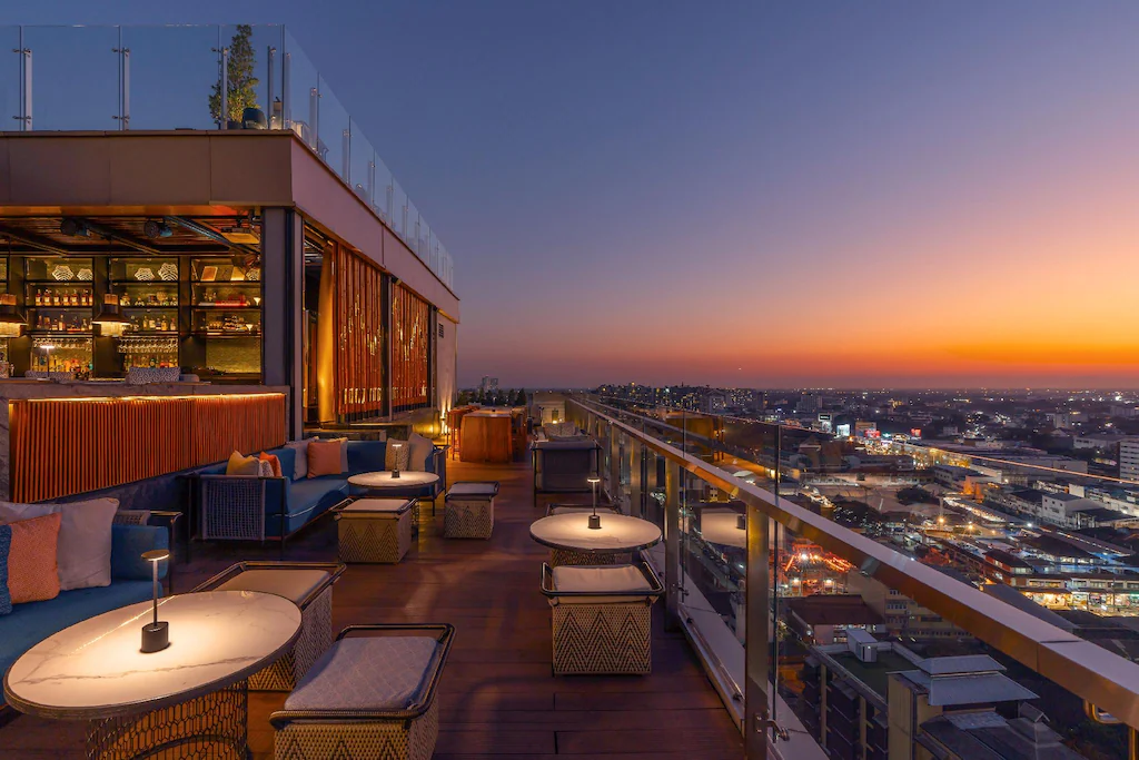 บาร์บนดาดฟ้าพร้อมที่นั่งเลานจ์และโต๊ะเล็ก ๆ มองเห็นทิวทัศน์เมืองยามพระอาทิตย์ตกดิน พร้อมแสงไฟที่ส่องประกายทั่วเมืองเบื้องล่าง—สถานที่ที่เหมาะอย่างยิ่งสำหรับแขกที่เข้าพักที่ โรงแรมเชียงใหม่ เพื่อผ่อนคลายและดื่มด่ำกับทัศนียภาพอันงดงาม.