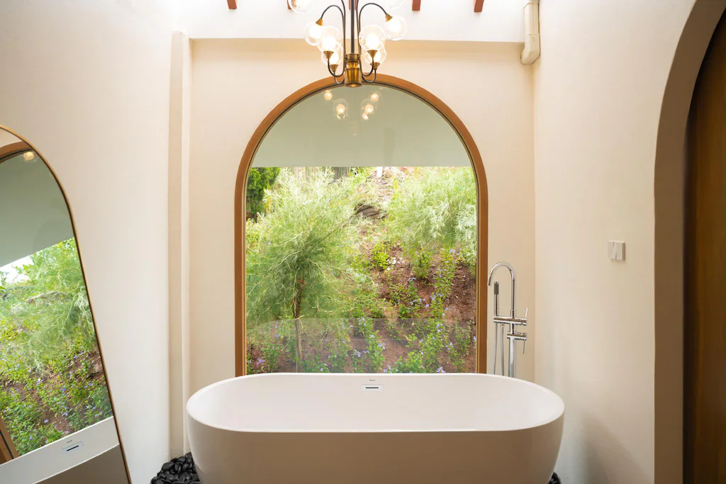 ห้องน้ำสไตล์มินิมอลที่ชวนให้นึกถึงโรงแรมในเชียงใหม่ โดดเด่นด้วยอ่างอาบน้ำสีขาวตั้งอยู่กลางห้อง ก่อนหน้าหน้าต่างโค้งขนาดใหญ่ที่มองเห็นวิวต้นไม้และธรรมชาติสีเขียว มีกระจกตั้งพื้นวางเอียงอยู่ทางซ้าย.