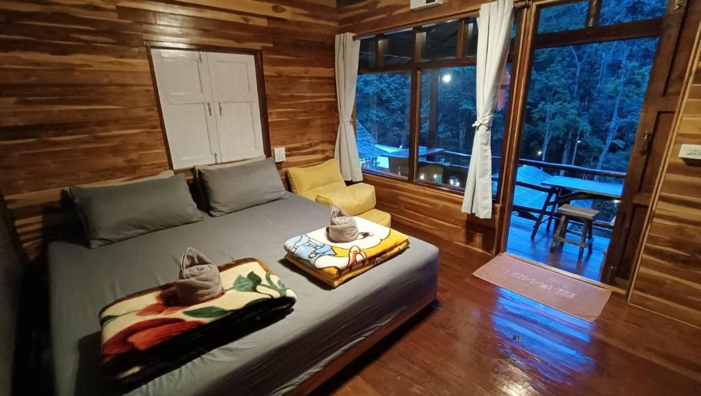 ห้องนอนในกระท่อมไม้ที่อบอุ่นในรีสอร์ทเชียงใหม่ พร้อมเตียงคู่ ผ้าเช็ดตัวพับไว้ ผ้าห่ม โซฟาสีเหลือง และประตูกระจกที่เปิดออกสู่ระเบียงพร้อมที่นั่งกลางแจ้งและวิวป่าที่เงียบสงบ.
