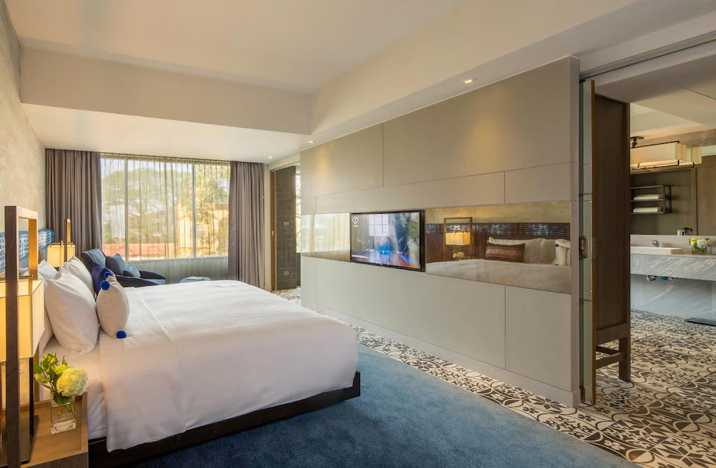 ห้องพักโรงแรมสมัยใหม่พร้อมเตียงขนาดใหญ่ ทีวีติดผนัง หน้าต่างบานใหญ่จากพื้นจรดเพดานพร้อมผ้าม่าน และวิวเปิดสู่ห้องน้ำที่มีพื้นกระเบื้องลายสวยงาม ณ โรงแรมเชียงใหม่สุดมีสไตล์แห่งนี้.